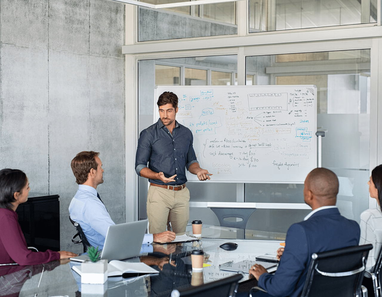 A man stands near a whiteboard with writing on it, presenting to four seated colleagues in a modern conference room. They are engaged, with laptops, notebooks, and a coffee cup on the glass table in front of them.